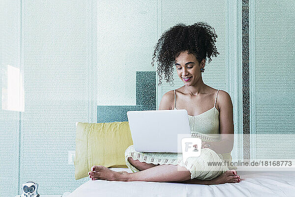 Smiling woman with curly hair using laptop on bed at home