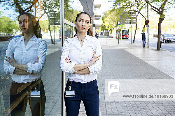 Confident businesswoman with arms crossed standing by glass on footpath