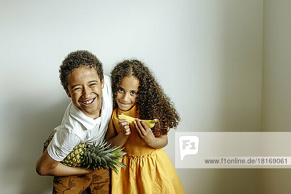 Happy brother with pineapple embracing sister holding banana in front of wall