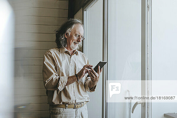 Smiling senior man standing near window using tablet PC at home