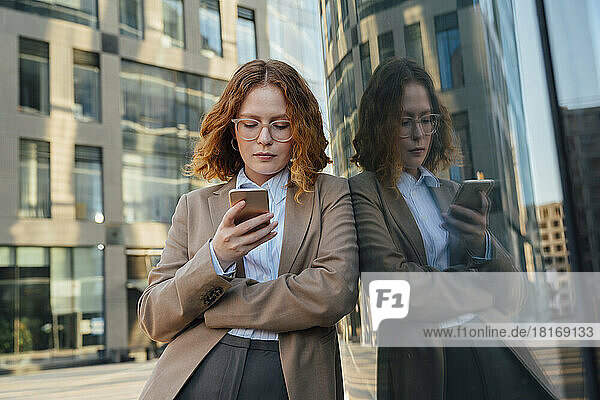 Businesswoman with eyeglasses using smart phone leaning on glass wall