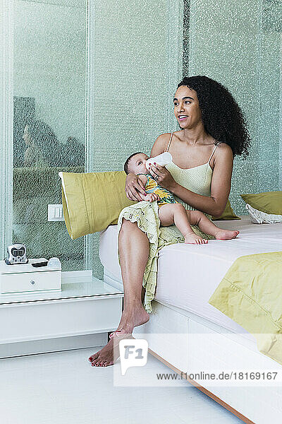 Happy woman feeding baby boy with bottle on bed at home