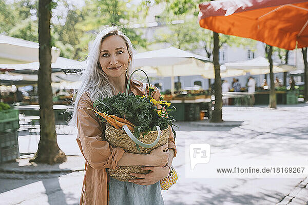 Smiling woman holding bag with vegetables and flower at market