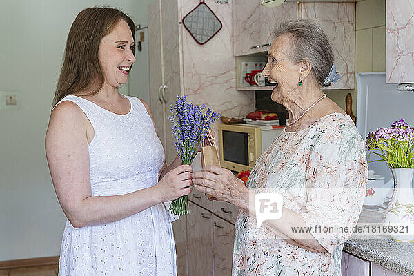 Smiling woman giving bunch of lavender flowers and gift to grandmother at home
