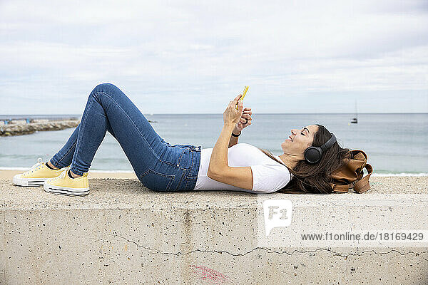 Happy woman lying on wall listening to music through wireless headphones and using smart phone