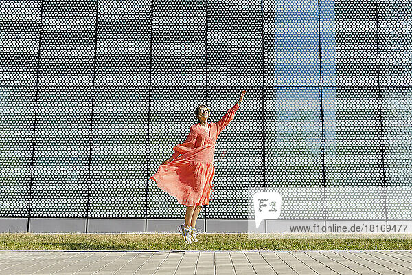 Glückliche Frau springt mit erhobener Hand auf Fußweg vor Mauer