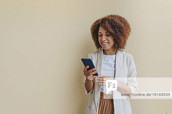 Smiling young businesswoman using mobile phone against beige background