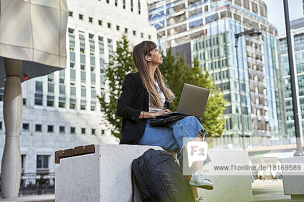Freelancer with laptop sitting on seat in front of buildings