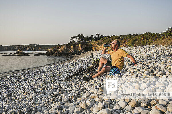 Man drinking water by bicycle sitting on pebbles at beach