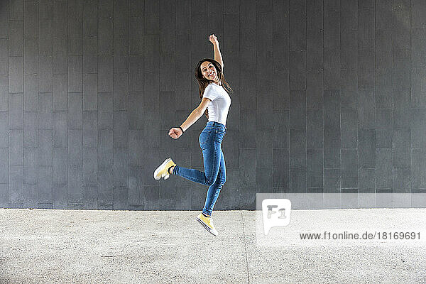 Happy woman with hand raised jumping on footpath in front of gray wall