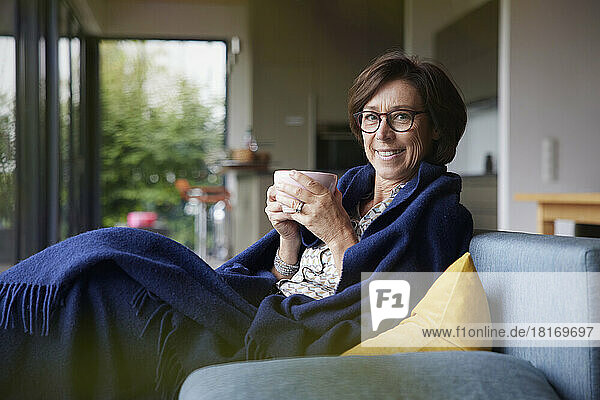 Glückliche Frau mit Kaffeetasse und Decke sitzt zu Hause auf dem Sofa