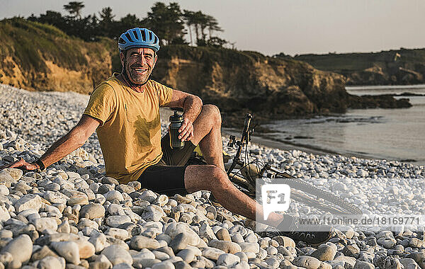 Happy man sitting by bicycle holding water bottle at beach