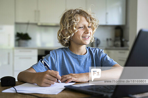 Fröhlicher Junge macht Hausaufgaben und schaut zu Hause auf den Laptop