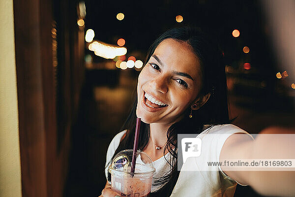 Cheerful woman with smoothie taking selfie at night