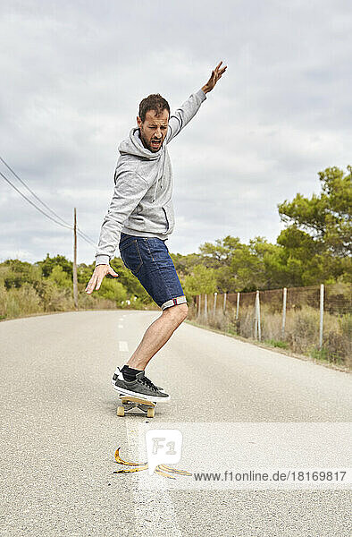 Terrified man with hand raised skateboarding by banana peel on road