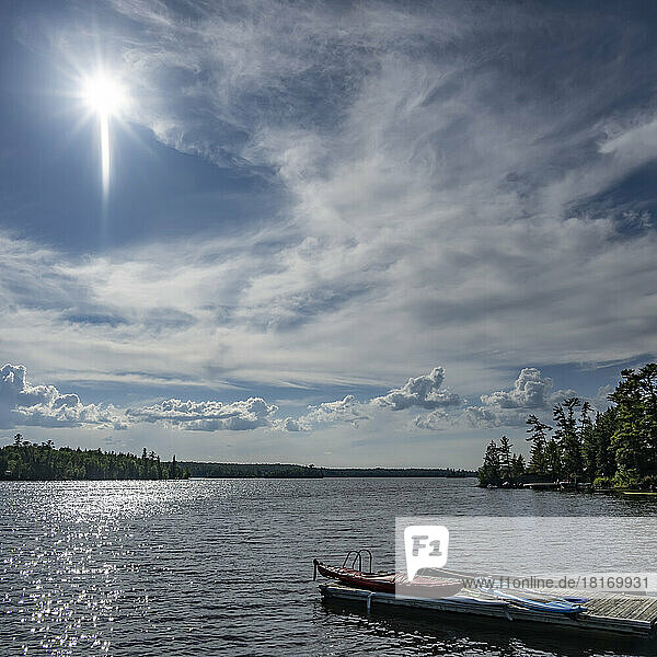 Das Sonnenlicht glitzert auf dem See im Cottage-Land  mit einem Kanu  einem Kajak und einem Paddelbrett auf dem Steg am Ufer  Lake of the Woods  Ontario; Ontario  Kanada