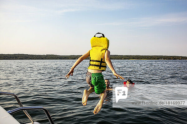Young boy jumping off a boat and swimming in a lake with a lifejacket and his father and brother watching him  Lac Ste. Anne; Alberta Beach  Alberta  Canada