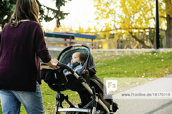 Eine Mutter geht mit ihrem Baby mit Down-Syndrom in einem Kinderwagen entlang eines Flusses in einem Stadtpark mit einer Brücke im Hintergrund während der Herbstsaison; St  Albert  Alberta  Kanada