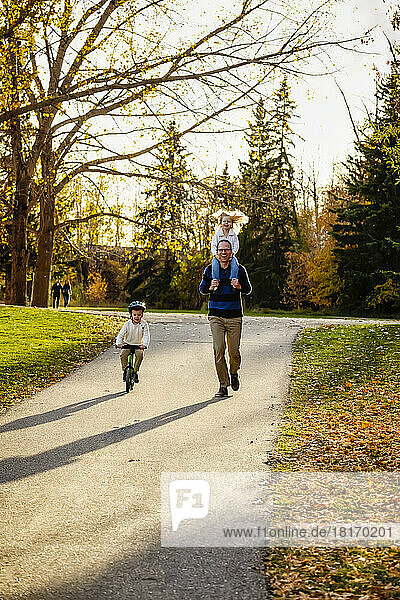 Ein Vater  der seine kleine Tochter auf den Schultern trägt  während er in einem Stadtpark spazieren geht  und sein kleiner Sohn  der neben ihnen mit dem Fahrrad fährt  während der Herbstsaison; St. Albert  Alberta  Kanada