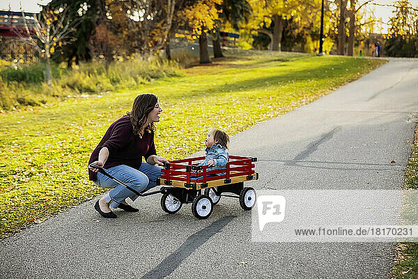 A mother pulling her baby with Down Syndrome in a wagon along a river in a city park with a bridge in the background during the fall season; St  Albert  Alberta  Canada