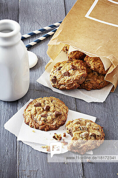 Milk bottle and oatmeal chocolate chip cookies in a brown paper bag and a cookie with a bite taken out of it with crumbs