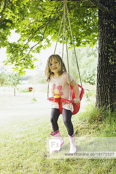 3 year old girl in rubber boots sitting in red swing in back yard  looking at camera and smiling  Sweden