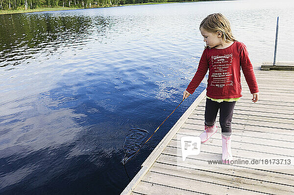 3 year old girl in red shirt on a pier holding a stick and playing in the water  Sweden