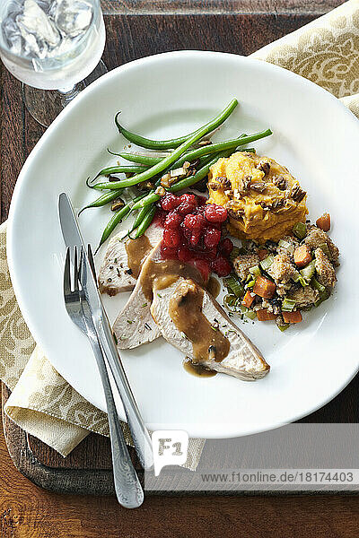 Overhead image of a holiday dinner plate containing turkey breast  gravy  stuffing  sweet potato  green beans  cranberries  walnuts and fresh rosemary. Cutlery  ice water  and a napkin are also present on a wooden cutting board on a table top.