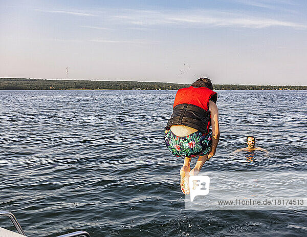 Young boy jumping off a boat and swimming in a lake with a lifejacket with his father watching from the water  Lac Ste. Anne; Alberta Beach  Alberta  Canada