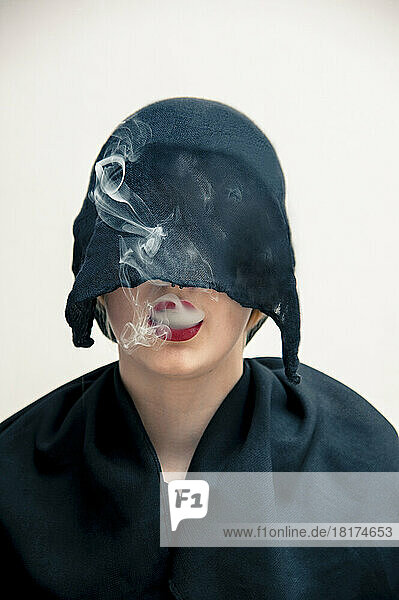 Close-up portrait of young woman wearing black  muslim dress and black  hijab covering part of head  while blowing smoke from red lips  studio shot on white background