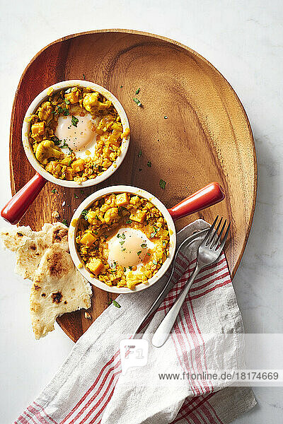 Two ramekins with lentil curry and baked eggs with naan bread on a wooden serving platter