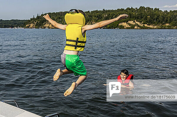 Young boy jumping off a boat and swimming in a lake with a lifejacket and his brother watching him  Lac Ste. Anne; Alberta Beach  Alberta  Canada