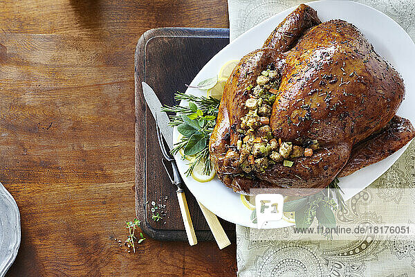 Holiday whole cooked turkey with glaze  herbs  and stuffing on a bed of fresh herbs and lemons. On a white serving platter with carving utensils on a wooden tabletop.
