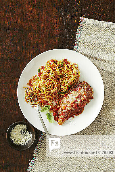 Plate of chicken parmesan with spaghetti and tomato sauce and a side dish with a fork and a side dish of cheese on a wooden background