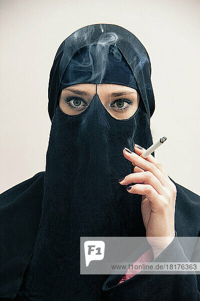 Close-up portrait of young woman wearing black  muslim hijab and muslim dress  holding cigarette and smoking  looking at camera  eyes showing eye makeup  studio shot on white background