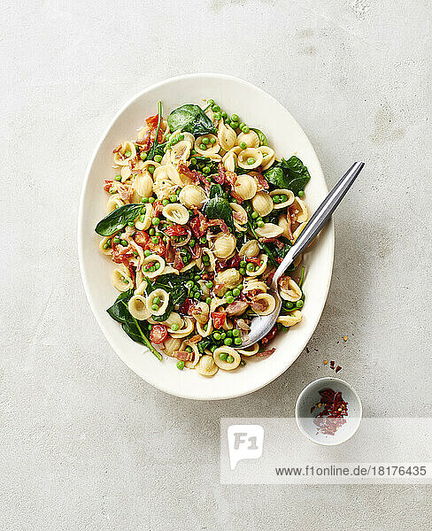 Platter of orecchiette pasta with peas  bacon  spinach and tomatoes and chili flakes in a small bowl on a beige background