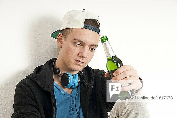 Close-up of teenage boy  sitting and leaning against wall  wearing hat and headphones around neck  holding bottle of beer  studio shot on white background
