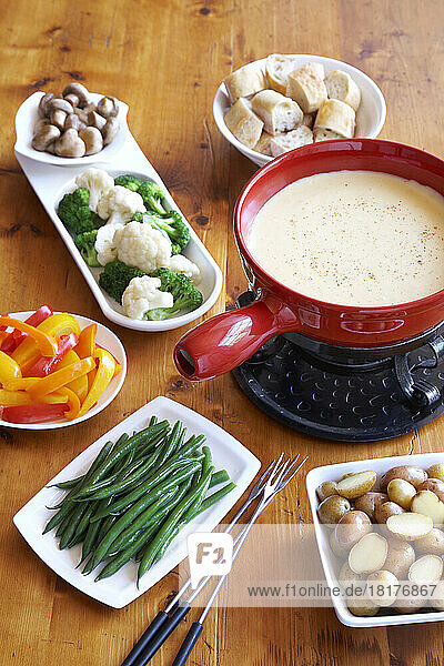 Cheese fondue in red pot with dishes of vegetables and bread on a wooden table