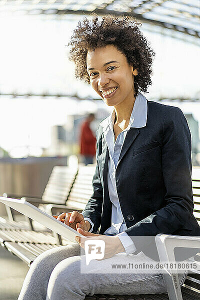 Happy businesswoman with tablet PC sitting on bench at railroad station