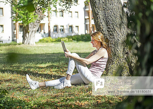 Lächelnde Frau mit Tablet-PC sitzt an einem sonnigen Tag am Baumstamm im Park