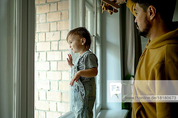 Netter Junge schaut durch das Fenster  während sein Vater zu Hause ein Flugzeugspielzeug in der Hand hält