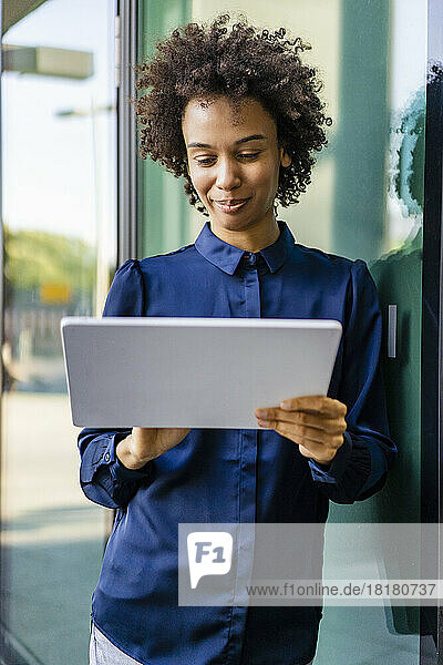Smiling businesswoman using tablet PC leaning on glass wall