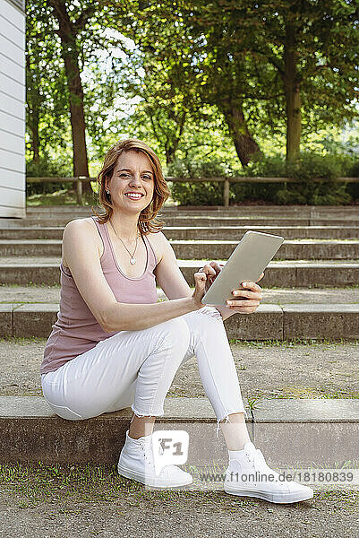 Glückliche Frau mit Tablet-PC sitzt auf der Treppe im Park