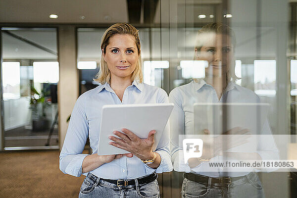 Smiling businesswoman with tablet PC leaning on glass wall at workplace