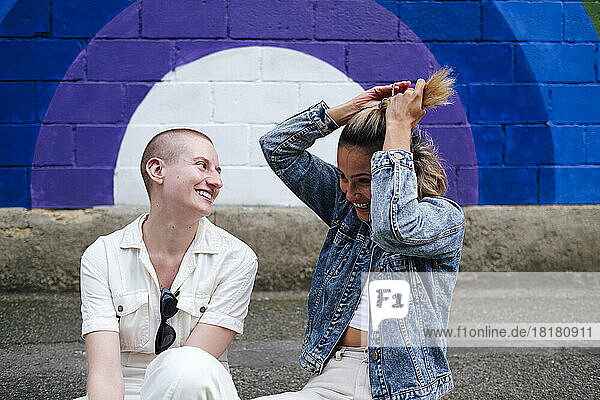 Happy woman tying hair with non-binary person sitting on footpath