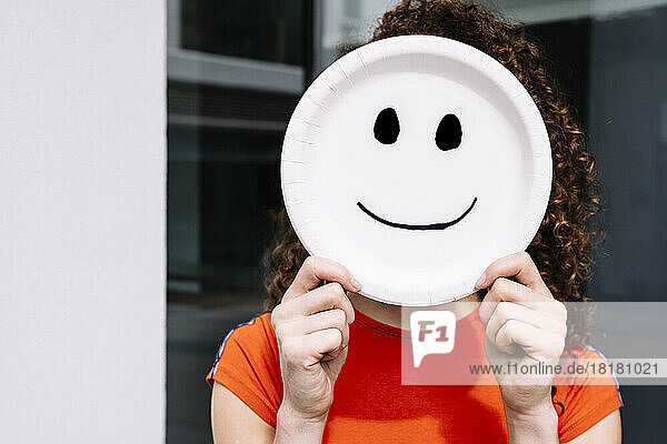 Young woman holding smiling emoticon plate over face