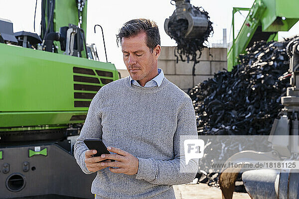 Mature businessman using smart phone in front of excavator