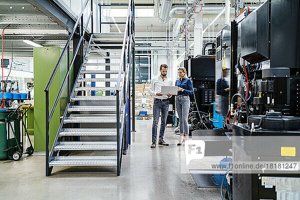 Business colleagues discussing over laptop standing by machinery in production hall at factory