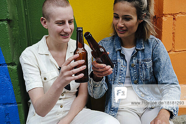 Schwules Paar toastet Bierflaschen und sitzt vor einer bunten Wand