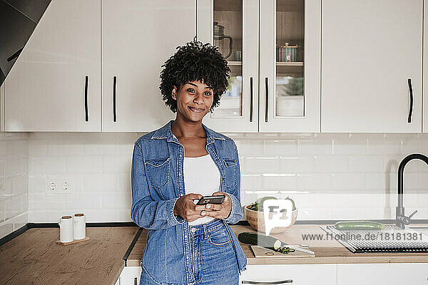 Smiling young woman with smart phone leaning on kitchen counter at home
